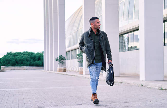 Personal masculino hispano feliz de cuerpo completo con maletín sonriendo y mirando hacia otro lado mientras camina sobre el pavimento cerca de un edificio moderno en la ciudad - foto de stock