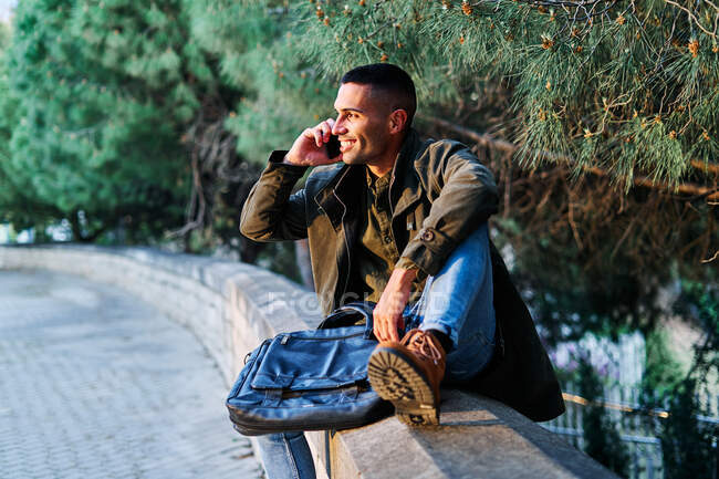 Homem hispânico otimista em roupas casuais sentado na fronteira perto da árvore conífera e olhando para o lado com sorriso ao ter uma conversa de smartphone no parque — Fotografia de Stock