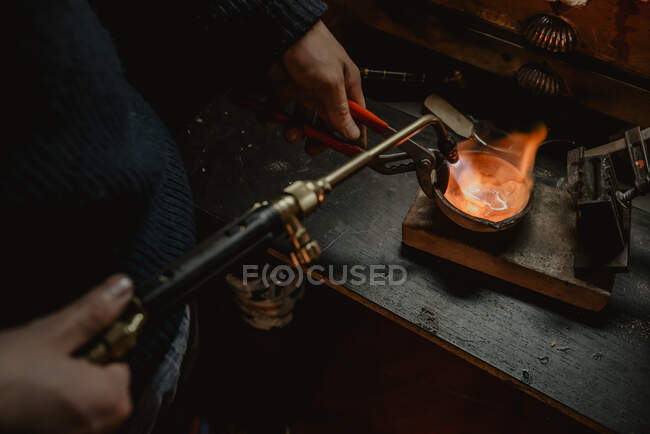 Неузнаваемый ювелир плавит металл для ювелирных изделий с паяльной лампой, стоя возле верстака в мастерской — стоковое фото