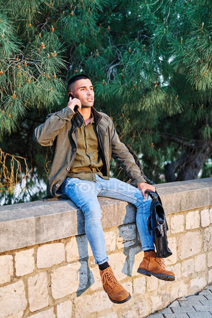 Tipo hispano con ropa casual mirando hacia otro lado y contestando una llamada telefónica mientras está parado cerca de la barrera y el árbol de coníferas en el parque - foto de stock