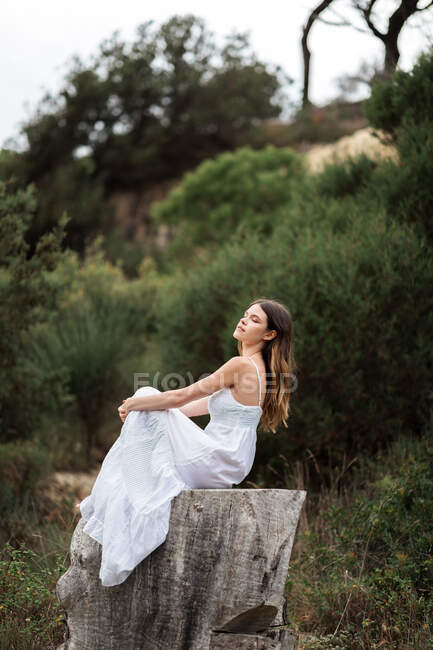 Side view of serene female in elegant white dress sitting on stump of tree in forest and embracing knees while enjoying nature with closed eyes — Stock Photo