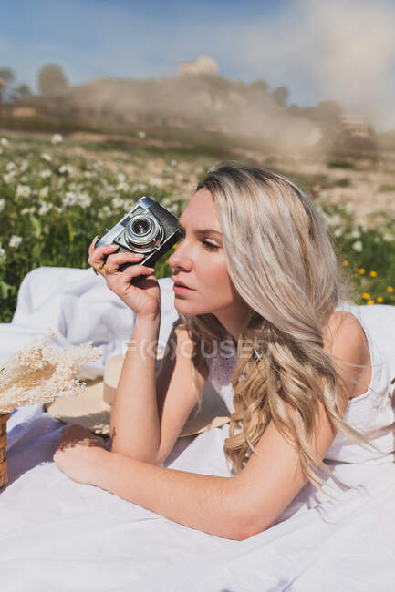 Mulher positiva deitada na xadrez e tirando foto na câmera à moda antiga no dia de verão no campo — Fotografia de Stock