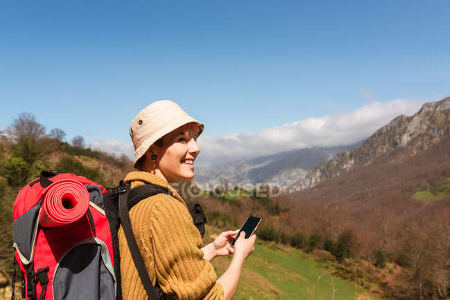 Vista laterale di felice zaino in spalla femminile utilizzando sullo smartphone mentre si gode l'avventura in estate sugli altopiani — Foto stock