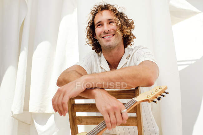 Músico masculino encantado com cabelo encaracolado sentado em cadeira de madeira com ukulele no fundo de cortinas brancas e olhando para longe — Fotografia de Stock