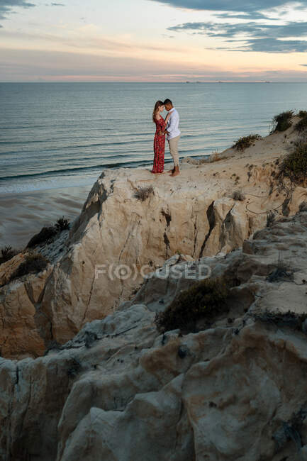 Amando casal multirracial em roupas elegantes abraçando na colina no fundo do céu por do sol sobre o mar no verão — Fotografia de Stock