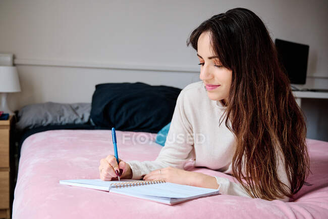 Estudiante acostada en la cama y escribiendo en bloc de notas mientras hace la tarea - foto de stock