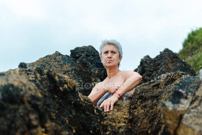 Low angle of aged gray haired bare shouldered female wrapped in towel looking away while smoking a cigarette on beach — Stock Photo