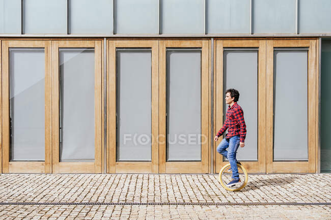 Vista laterale del corpo pieno di giovane maschio attivo in camicia a scacchi e jeans che eseguono trucco su monociclo vicino alla parete di vetro specchiato dell'edificio contemporaneo — Foto stock