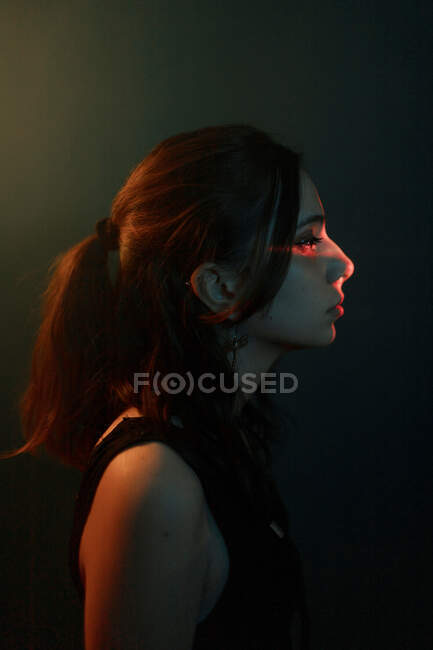 Seitenansicht des jungen weiblichen Modells mit Lichtprojektion auf das Gesicht, das im dunklen Studio steht und wegschaut — Stockfoto