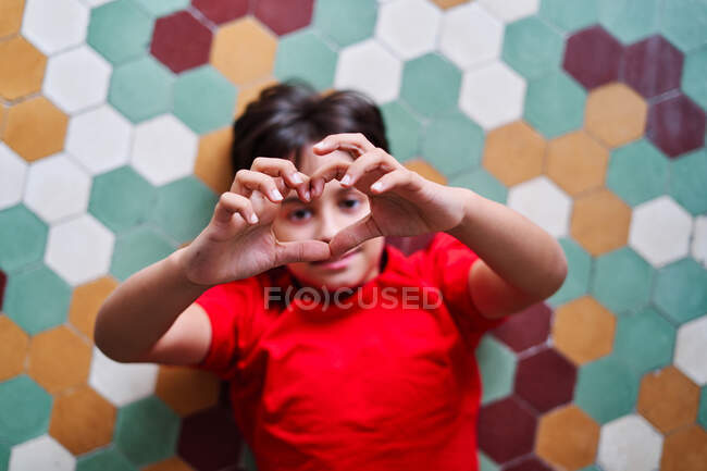 From above of preteen boy lying on floor and making heart shaped sign with fingers while looking at camera — Stock Photo