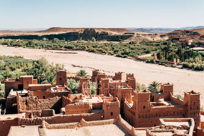 Edifícios antigos de pedra marrom entre plantas tropicais verdes e deserto com céu azul claro no fundo em Marrocos — Fotografia de Stock