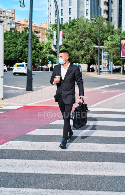 Cuerpo completo de empresario masculino joven bien vestido en traje elegante y máscara protectora que lleva el maletín y la taza desechable de bebida para llevar caminando en el paso de peatones en el día de verano en la ciudad - foto de stock