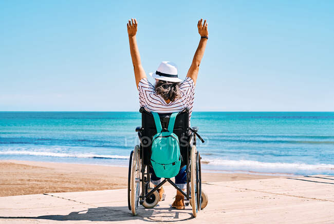 Вид ззаду на невпізнавану жінку-мандрівника в інвалідному візку з рюкзаком піднімаючи руки, насолоджуючись літньою подорожжю на пляжі біля блакитного моря — стокове фото