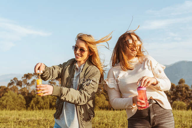Enge Freundinnen mit Sonnenbrille pusten gemeinsam Seifenblasen auf einer Wiese in den Bergen — Stockfoto