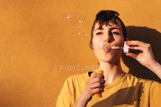 Mulher moderna com piercing soprando bolhas de sabão no dia ensolarado contra a parede amarela — Fotografia de Stock