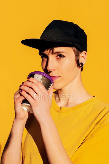 Jeune femme informelle dans le chapeau à la mode sirotant à emporter boisson chaude sur fond jaune — Photo de stock