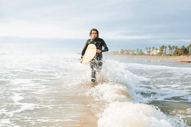 Мужчина-серфер, одетый в гидрокостюм, бегает с доской для серфинга на пляже с восходом солнца — стоковое фото