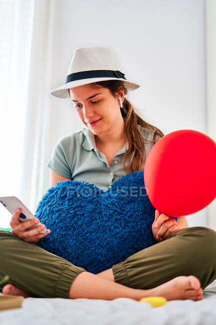 Mujer joven con ropa casual y sombrero sosteniendo globo rojo y cojín en forma de corazón navegando en el teléfono inteligente mientras celebra su cumpleaños sola en la cama con comida y jugo - foto de stock