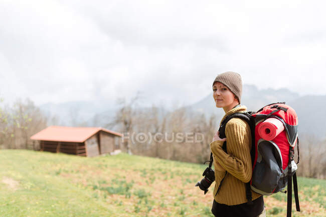 Вид сбоку на застенчивую женщину с рюкзаком, гуляющую в горах, глядя в камеру — стоковое фото