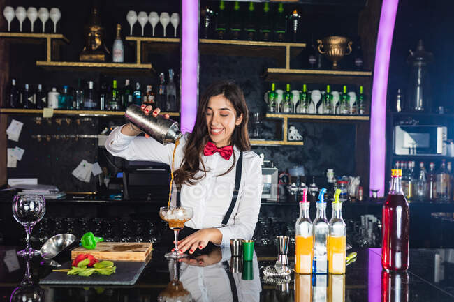Cantinera sonriente con elegante atuendo que sirve cóctel de una coctelera en un vaso mientras está de pie en el mostrador en el bar moderno - foto de stock