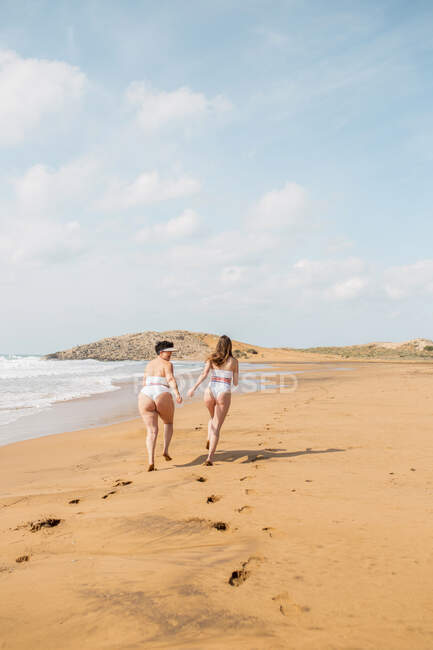 Visão traseira de amigas felizes em trajes de banho correndo no oceano espumoso perto da praia de areia sob o céu azul nublado em dia ensolarado — Fotografia de Stock