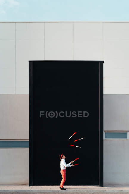 Seitenansicht eines nicht wiederzuerkennenden männlichen Zirkusjongleurs, der einen Trick mit Jonglierkeulen gegen die hohe schwarze Wand eines modernen Gebäudes aufführt — Stockfoto