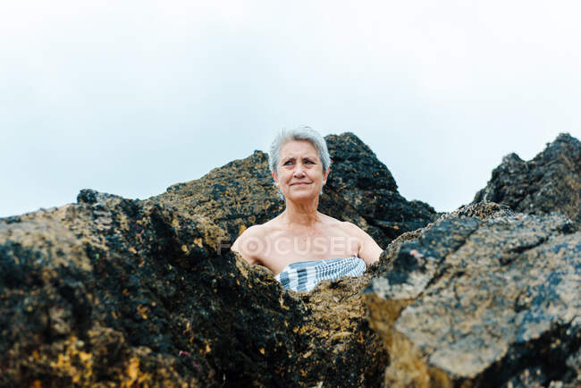Low angle of aged gray haired bare shouldered female wrapped in towel looking away while hiding amidst large rocky boulders on beach — Stock Photo