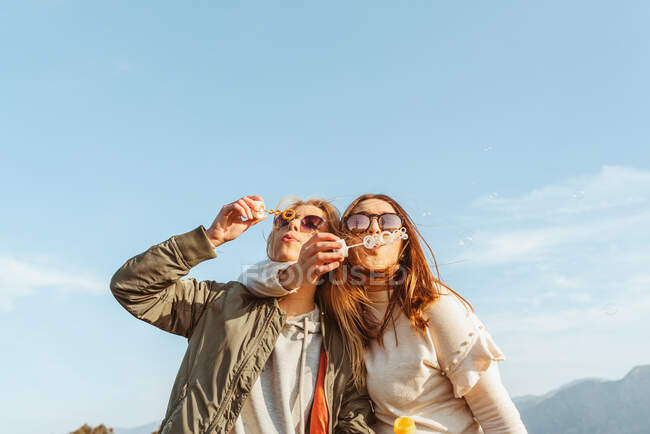 Fechar amigos do sexo feminino em óculos de sol soprando bolhas de sabão juntos de pé no abraço no prado nas montanhas — Fotografia de Stock
