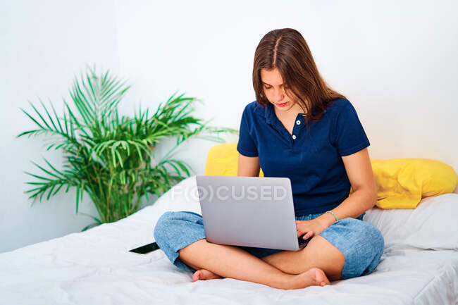 Позитивная молодая женщина-фрилансер в повседневной одежде сидит со скрещенными ногами на кровати и печатает на ноутбуке, работая над удаленным проектом дома — стоковое фото