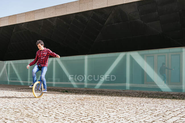 Full body side view of active young male in checkered shirt and jeans performing trick on unicycle near mirrored glass wall of contemporary building — Stock Photo