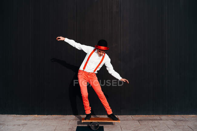 Corps complet d'artiste de cirque masculin en pantalon rouge et chapeau performant tour sur le tableau d'équilibre contre le mur noir — Photo de stock