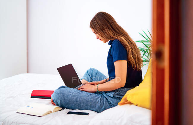 Вид сбоку позитивной молодой женщины-фрилансера в повседневной одежде, сидящей со скрещенными ногами на кровати и сидящей на ноутбуке, работая на удаленном проекте дома — стоковое фото
