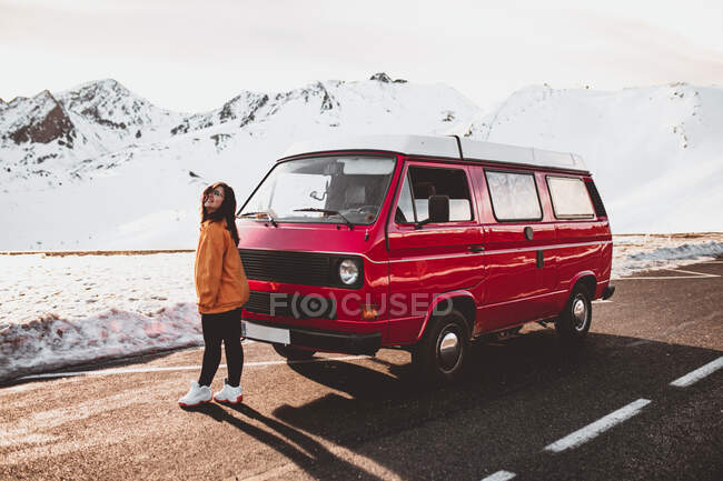 Jovem turista feliz em óculos de pé perto de automóvel vintage entre terreno deserto na neve perto de montanhas — Fotografia de Stock