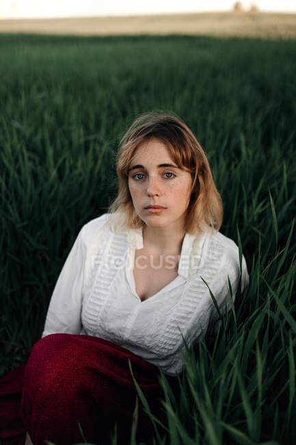 Peaceful young female in retro styled white blouse sitting amidst tall green grass and looking at camera while resting in summer evening in countryside — Stock Photo