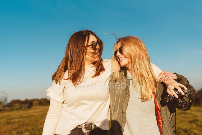 Молодые улыбающиеся девушки с фотокамерой, смотрящие друг на друга и обнимающиеся с голубым небом в поле — стоковое фото