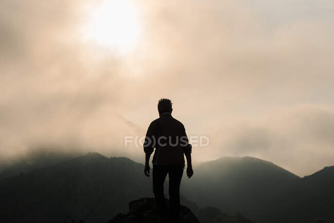 Silhouette of anonymous explorer admiring mountainous terrain against cloudy sunrise sky in morning in nature — Stock Photo