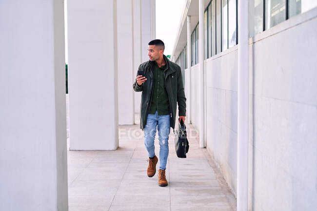 Ganzkörperhispanischer Mann in lässiger Kleidung, der wegschaut und sein Handy in der Hand hält, während er vor der Arbeit auf dem Bürgersteig steht — Stockfoto