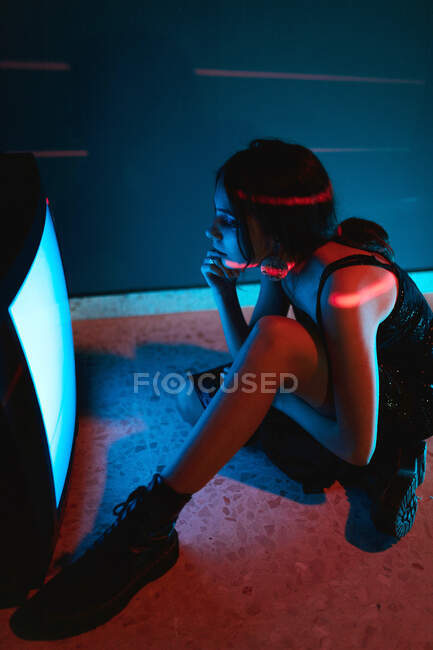 Side view of unrecognizable female model in black dress sitting on floor near glowing old television in dark studio — Stock Photo