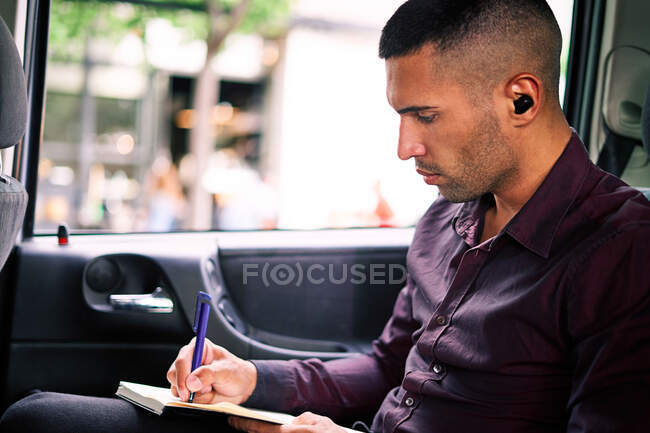 Homem empreendedor hispânico focado sentado no assento do passageiro do carro e escrevendo no planejador enquanto viaja para o trabalho — Fotografia de Stock