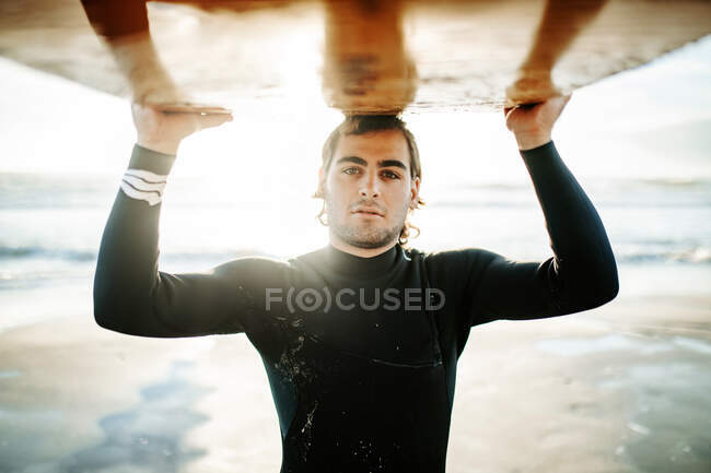 Портрет молодого серфінгу, одягненого в гідрокостюм, стоїть на камеру на пляжі з дошкою для серфінгу над головою під час сходу сонця — стокове фото