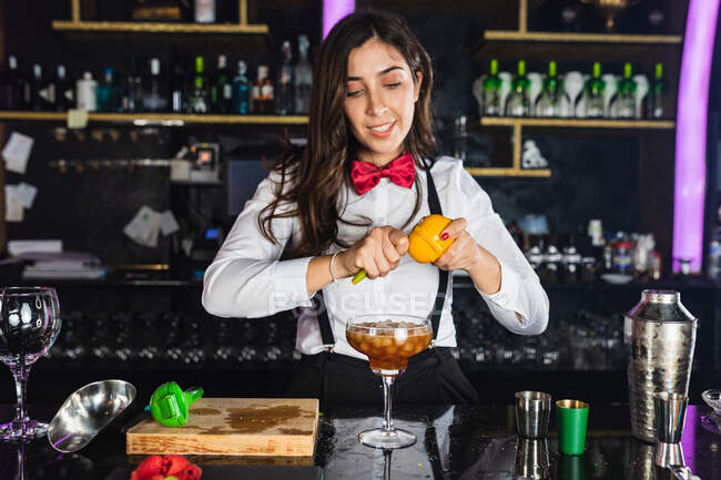 Young female barkeeper in stylish outfit decorating cocktail with lemon peel while standing at counter in modern bar — Stock Photo