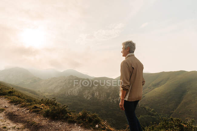Blick zurück auf die stehende Seniorin, die das bergige Gelände vor wolkenverhangenem Himmel am Morgen in der Natur bewundert — Stockfoto
