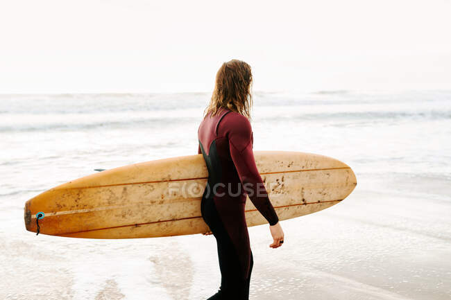 Vista laterale dell'uomo surfista vestito di muta che cammina guardando lontano con la tavola da surf verso l'acqua per catturare un'onda sulla spiaggia durante l'alba — Foto stock