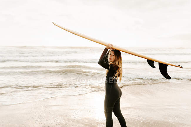 Vista lateral da surfista vestida de fato de mergulho em pé olhando para a câmera enquanto segurava a prancha na cabeça na praia durante o nascer do sol no fundo — Fotografia de Stock