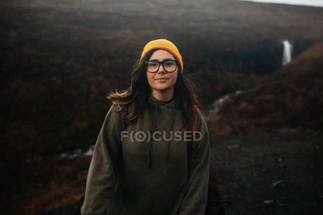 Jovem turista em óculos e chapéu com piercing olhando para a câmera na colina perto de cachoeira e rio de montanha — Fotografia de Stock