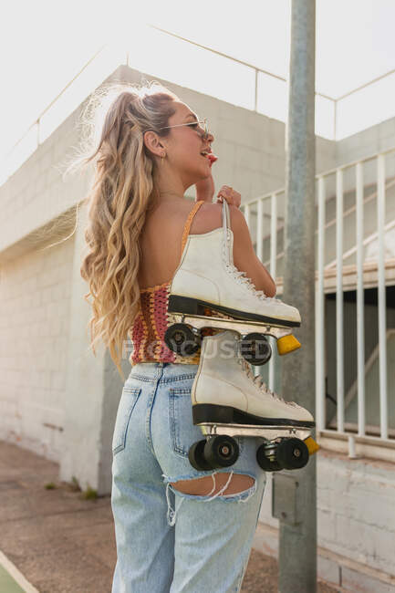 Visão traseira atraente jovem fêmea em jeans rasgados carregando patinadores no ombro e de pé na rua — Fotografia de Stock