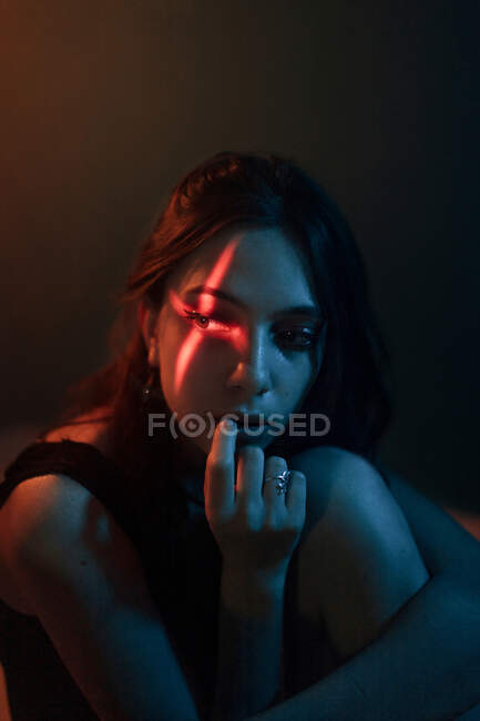 Молодая неэмоциональная женщина-модель с крестообразной светлой проекцией на лице сидит в темной студии и смотрит в сторону — стоковое фото