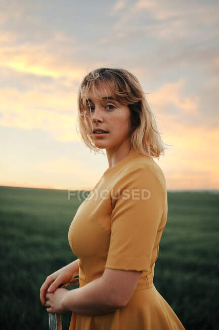Side view of female in vintage style dress standing on ladder in green grassy field against cloudy sunset sky and looking at camera as concept of dream and freedom — Stock Photo