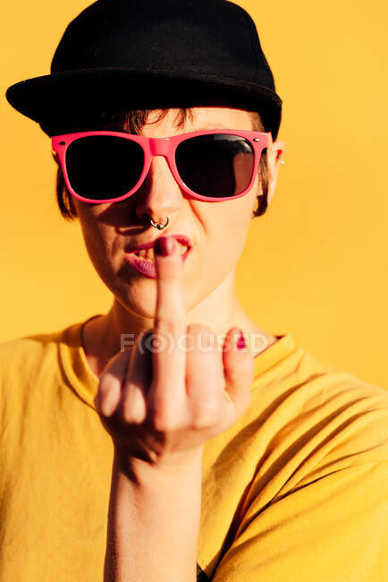 Rebellious female in black hip hop cap and sunglasses showing middle finger to camera against yellow background — Stock Photo