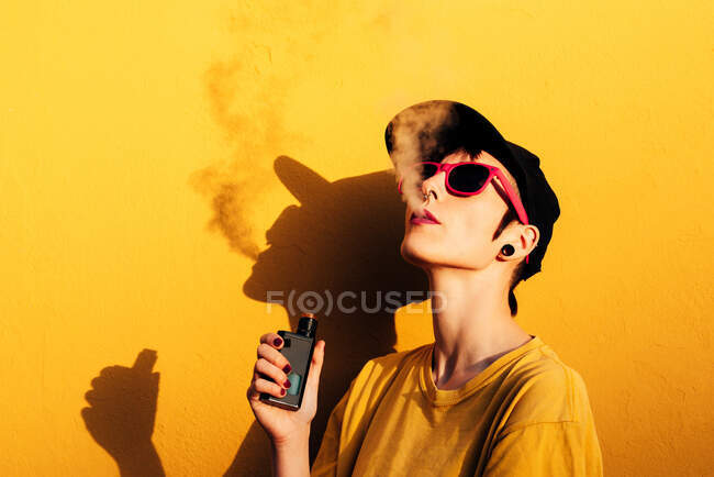 Mujer contemporánea en traje elegante exhalando humo mientras está de pie cerca de la pared amarilla y vapeando en la calle de la ciudad - foto de stock